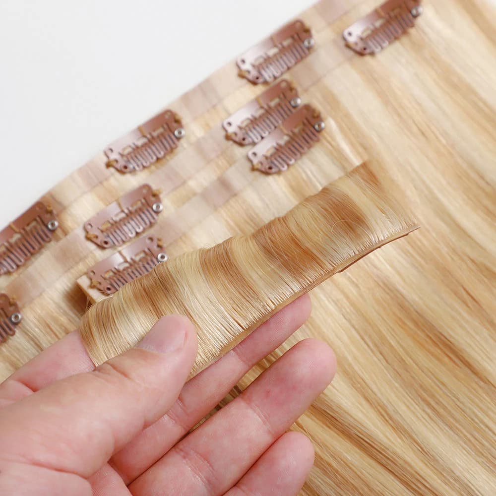 A hand lifting a blonde clip-in base edge above other pieces with metal clips visible