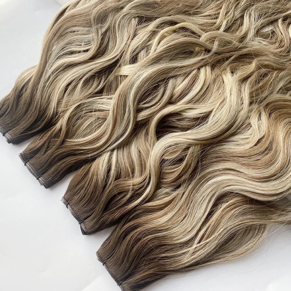 Wavy blonde and brown weft hair extensions arranged diagonally on a white surface